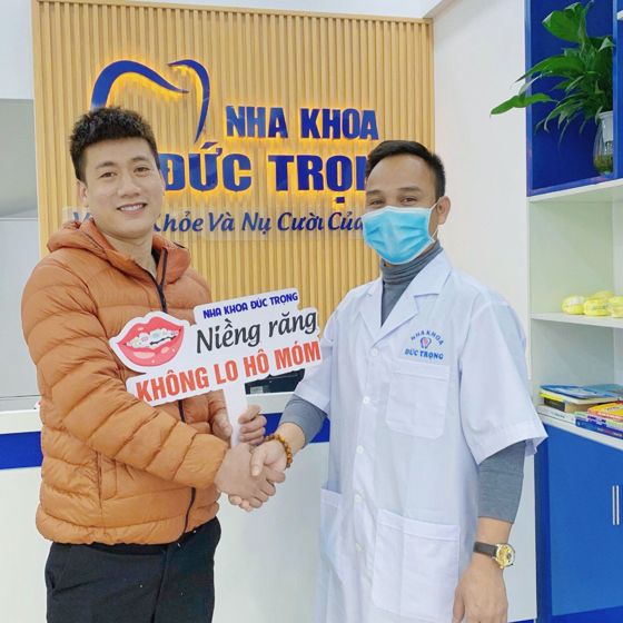 Khách hàng nam sau khi tháo mắc cài bắt tay bác sĩ tại quầy lễ tân, cầm biển Niềng răng không lo hô móm